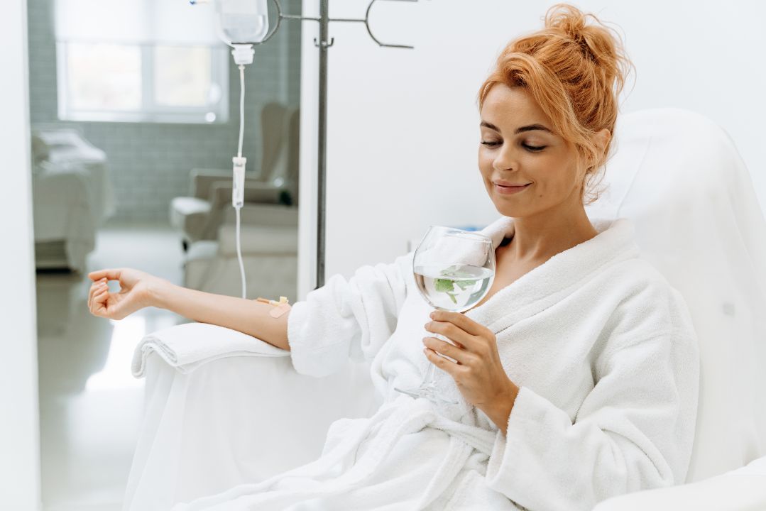 a charming woman in white bathrobe sitting in armchair and receiving IV infusion. She is holding glass of lemon beverage and smiling 