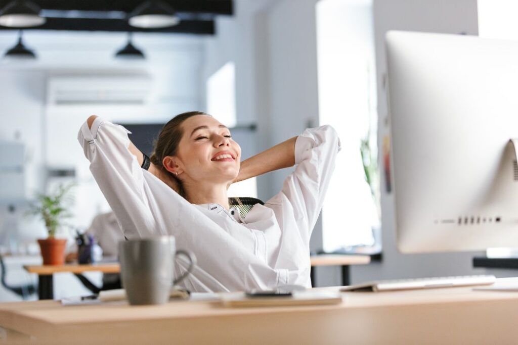 Happy young woman dressed in shirt sitting at her workplace at the office, resting, stretching her hands