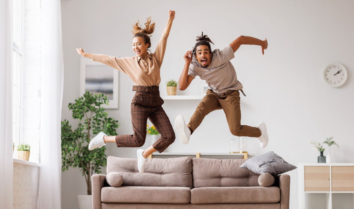 Full length of young energetic active african american family couple jumping on sofa in cozy living room, carefree black man and woman have fun together at home. Freedom and happiness in relationship