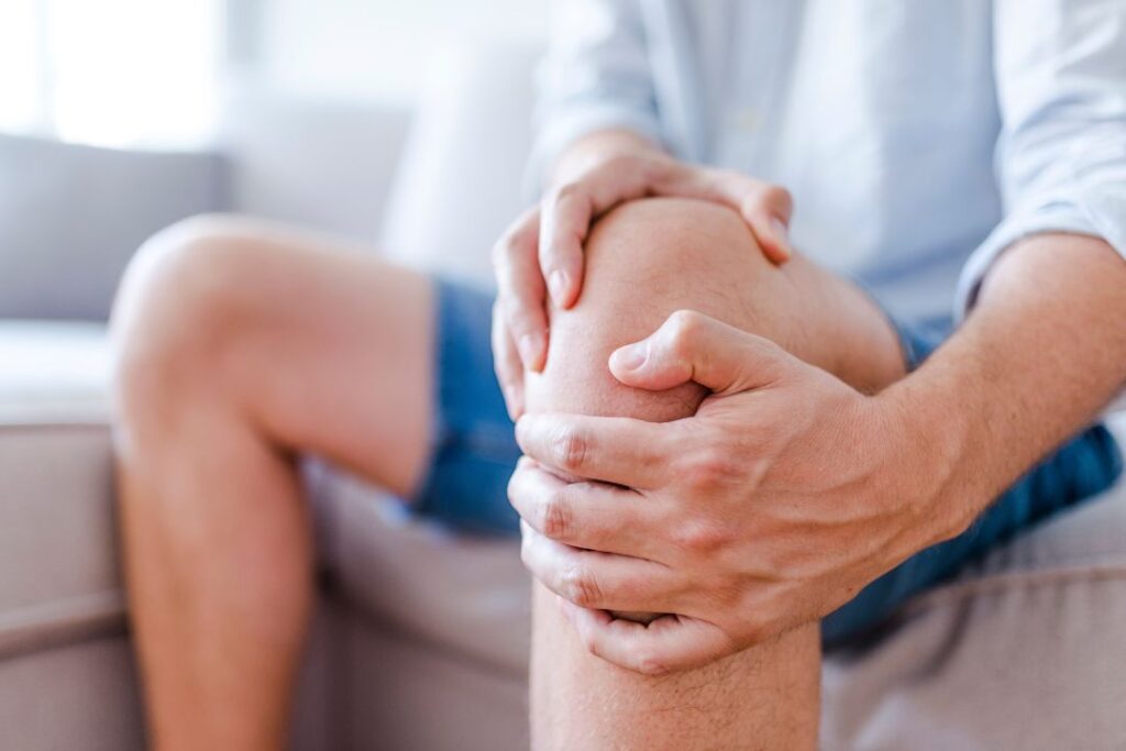 Man suffering from knee pain sitting sofa. A mature man massaging his painful knee. Man suffering from knee pain at home