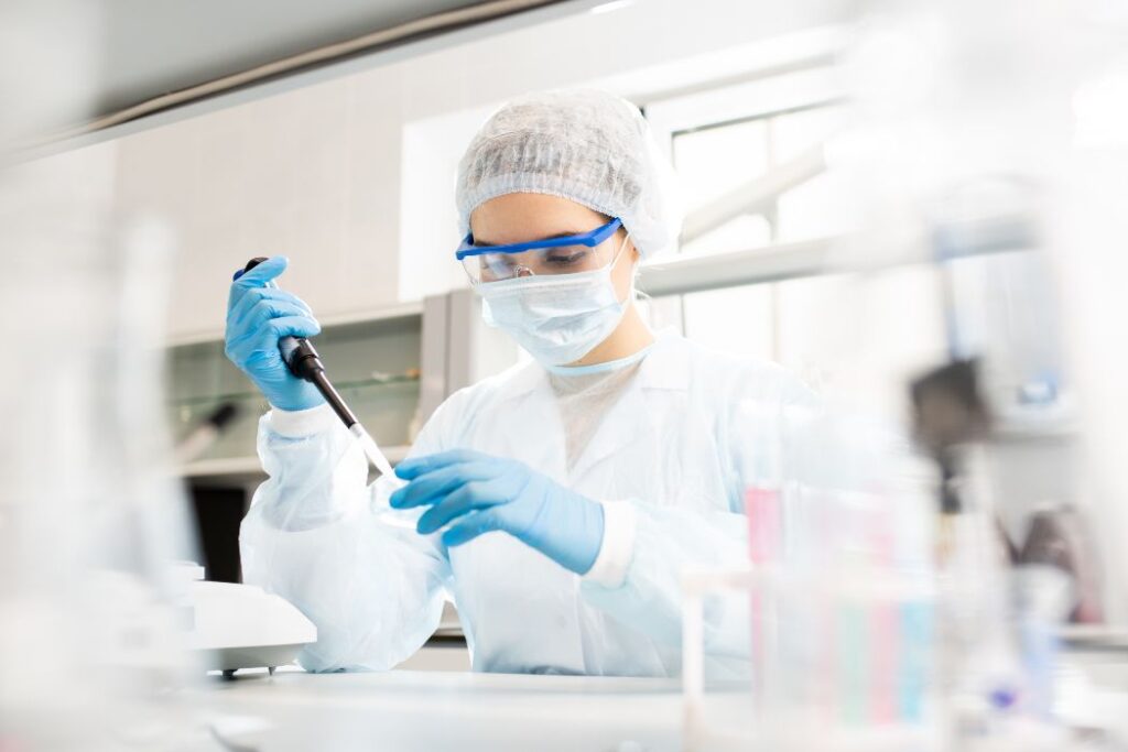 female microbiologist in sterile clothing and safety goggles sitting at table and dropping reagent in petri dish while doing research in laboratory 